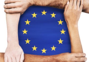 Multiracial group of hands and arms of men and women clasping each other, signifying unity, solidarity, cooperation and teamwork, around the EU flag.