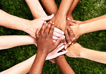 A pile of  multiracial hands are stacked in support or unity, against a background of grass. All for one and one for all!